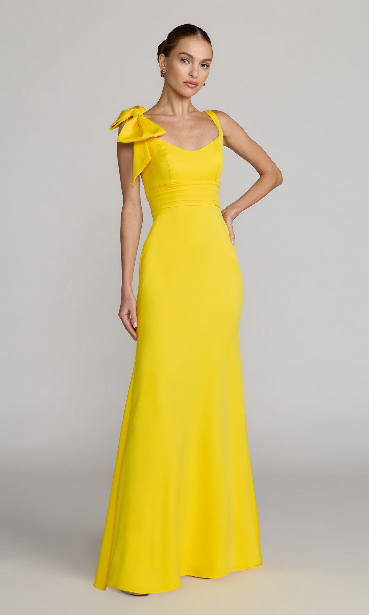 A woman wearing the Long Formal Dress: Mac Duggal 49964 in lemon—a crepe column gown featuring a large bow on one shoulder—stands against a neutral background.